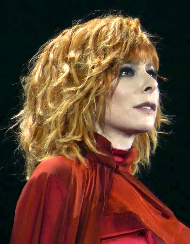 > "Portrait d’une artiste variété française en tenue rouge vif sur fond noir, cheveux blonds bouclés et regard de trois-quarts – visuel contrasté Vibes Concert"