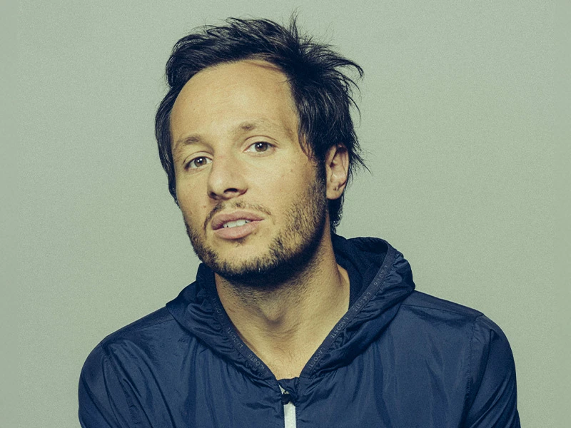 > "Portrait d’un artiste variété française en veste à capuche bleu marine sur fond gris clair, cheveux noirs décoiffés – esthétique sobre Vibes Concert"