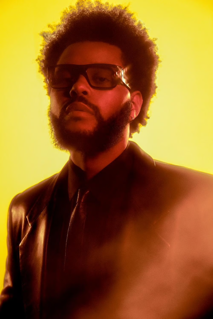 > "Portrait d’un artiste variété internationale en veste sombre et cravate sur fond jaune vif, cheveux bouclés volumineux – contraste visuel Vibes Concert"